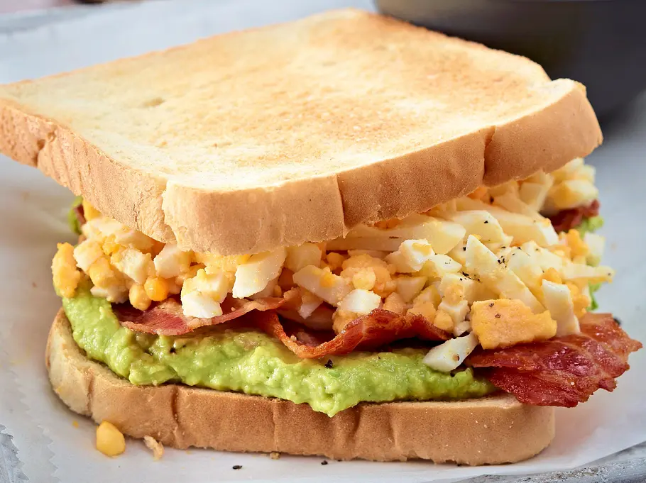 Knusprig getoastetes Sandwich mit cremigem Avocado-Mus, saftigem Rührei und kross gebratenem Bacon, serviert auf einem Backblech, dazu eine Schale frische Avocadocreme.