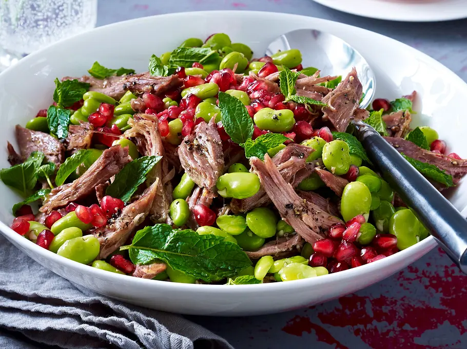 Beef-Bohnen-Salat Rezept Bunter Beef-Bohnen-Salat mit zartem Rindfleisch, grünen Bohnen, Granatapfelkernen und frischem Minzblatt in einer weißen Schale, daneben ein silbernes Besteck und eine graue Stoffserviette.