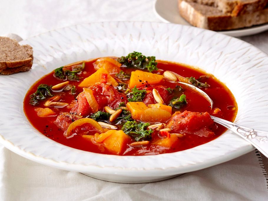 Kräftiger Eintopf mit Süßkartoffeln, Tomaten, Grünkohl und gerösteten Kernen in einem tiefen weißen Teller.
