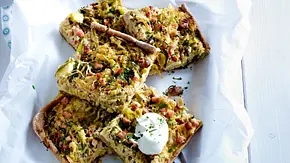 Herzhafter Brotkuchen mit Kasselerwürfeln und Sauerkraut, in Stücke geschnitten, auf einem mit Backpapier belegten Tablett, garniert mit einem Klecks Sauerrahm, darunter ein gemustertes Tuch auf heller Holzoberfläche. - Foto: LECKER @ Bauer Media Group
