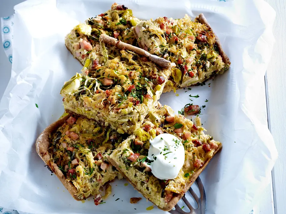 Herzhafter Brotkuchen mit Kasselerwürfeln und Sauerkraut, in Stücke geschnitten, auf einem mit Backpapier belegten Tablett, garniert mit einem Klecks Sauerrahm, darunter ein gemustertes Tuch auf heller Holzoberfläche.