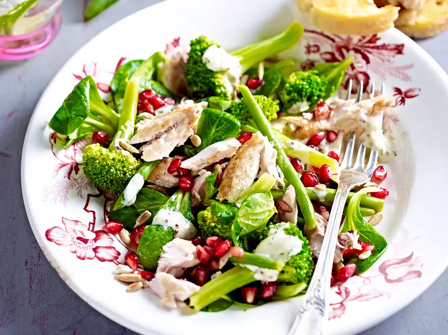 Leichter Salat mit Makrele und grünem Gemüse Frischer grüner Salat mit Brokkoli, Makrelenfilets, Granatapfelkernen und Kernen auf einem verzierten weißen Teller, dazu geröstetes Brot.