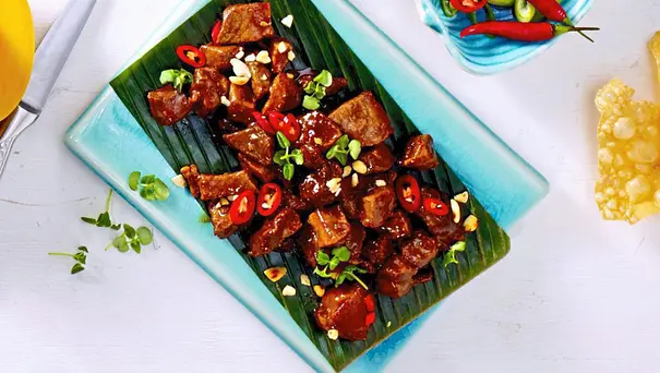 Würfel von karamellisiertem Schweinefleisch mit Chili und Frühlingszwiebeln, serviert auf einem grünen Teller und mit gehackten Erdnüssen bestreut. - Foto: LECKER @ Bauer Media Group