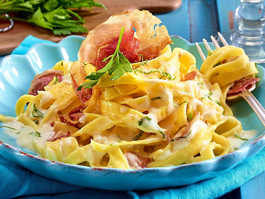 Bandnudeln in einer cremigen Limettenrahmsoße, serviert mit knusprigem Schinken und frischen Kräutern auf einem pastellblauen Teller.