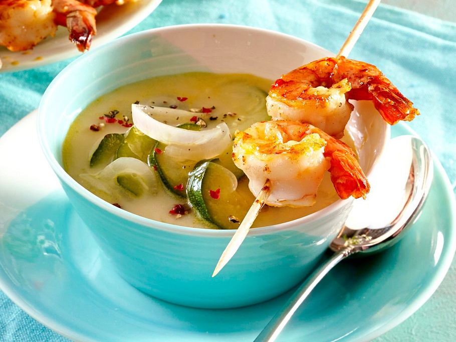 Cremige Zitronen-Joghurt-Suppe mit Zucchinischeiben und Zwiebeln, serviert mit gegrilltem Garnelenspieß.