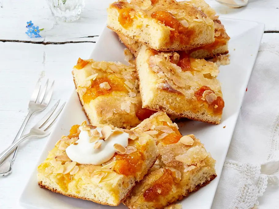 Saftiger Butterkuchen in rechteckigen Stücken auf einer weißen Platte, belegt mit glasierten Aprikosen und gehobelten Mandeln, zum Teil mit einem Klecks Sahne garniert.