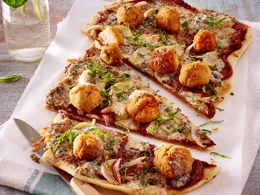Dünner, knuspriger Flammkuchen mit leuchtender Rote-Bete-Creme, cremigem Käse, zarten Zwiebelringen und goldbraun gebackenen Kichererbsenbällchen, frisch angerichtet auf Backpapier und einem Holzbrett. Frische Kräuter sorgen für einen grünen Farbakzent.