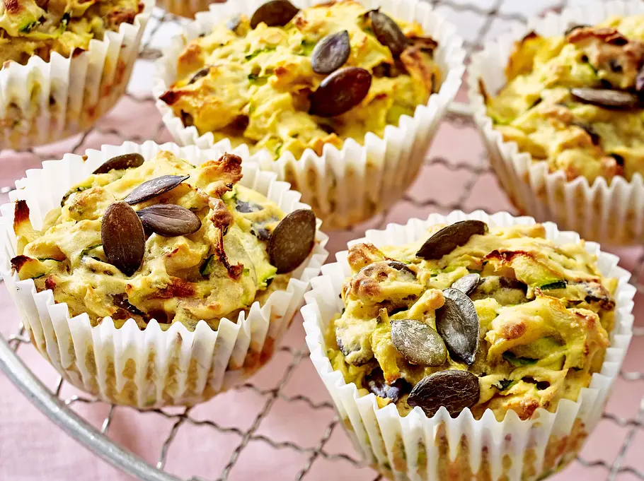 Saftige Zucchini-Muffins ohne Zuckerzusatz, bestreut mit Kürbiskernen, in weißen Papierförmchen auf einem runden Kuchengitter.