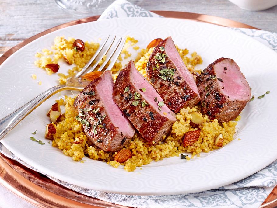 Zart rosa gebratenes Lammfilet, serviert auf aromatischem Gewürz-Couscous mit Mandeln und Kräutern auf einem weißen Teller.