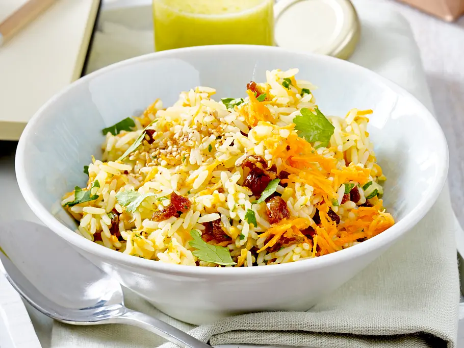 Orientalischer Reissalat mit Möhrenraspeln, Kräutern und Sesam in einer weißen Schale, dazu großer Löffel auf heller Serviette.