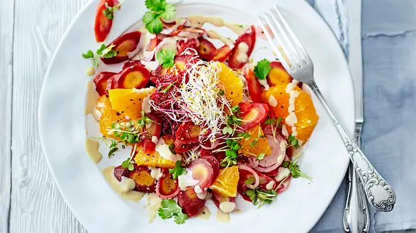 Bunter Salat aus fein geschnittenen Möhren und saftigen Orangen-Stücken, verfeinert mit frischem Koriander, Sprossen und cremigem Tahin-Dressing, auf einem weißen Teller angerichtet und von elegantem Besteck begleitet. - Foto: LECKER @ Bauer Media Group