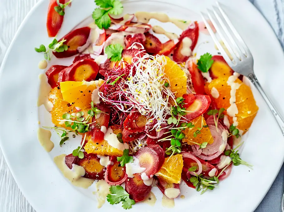 Bunter Salat aus fein geschnittenen Möhren und saftigen Orangen-Stücken, verfeinert mit frischem Koriander, Sprossen und cremigem Tahin-Dressing, auf einem weißen Teller angerichtet und von elegantem Besteck begleitet.
