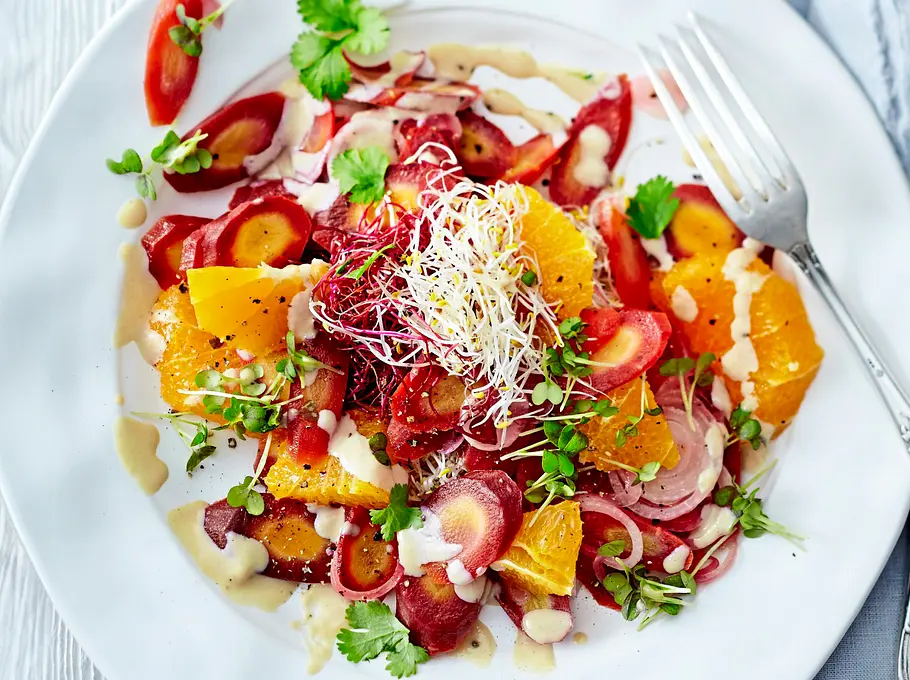 Bunter Salat aus fein geschnittenen Möhren und saftigen Orangen-Stücken, verfeinert mit frischem Koriander, Sprossen und cremigem Tahin-Dressing, auf einem weißen Teller angerichtet und von elegantem Besteck begleitet.