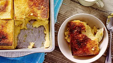 Goldbraun gebackener French-Toast-Auflauf in einer rechteckigen Form, mit knuspriger Oberfläche und saftiger Krume. Daneben ein Kännchen Vanillesoße und ein Stück des Auflaufs in einer Schale, serviert auf einem Holztisch mit Pfannenwender und Tuch. - Foto: LECKER @ Bauer Media Group