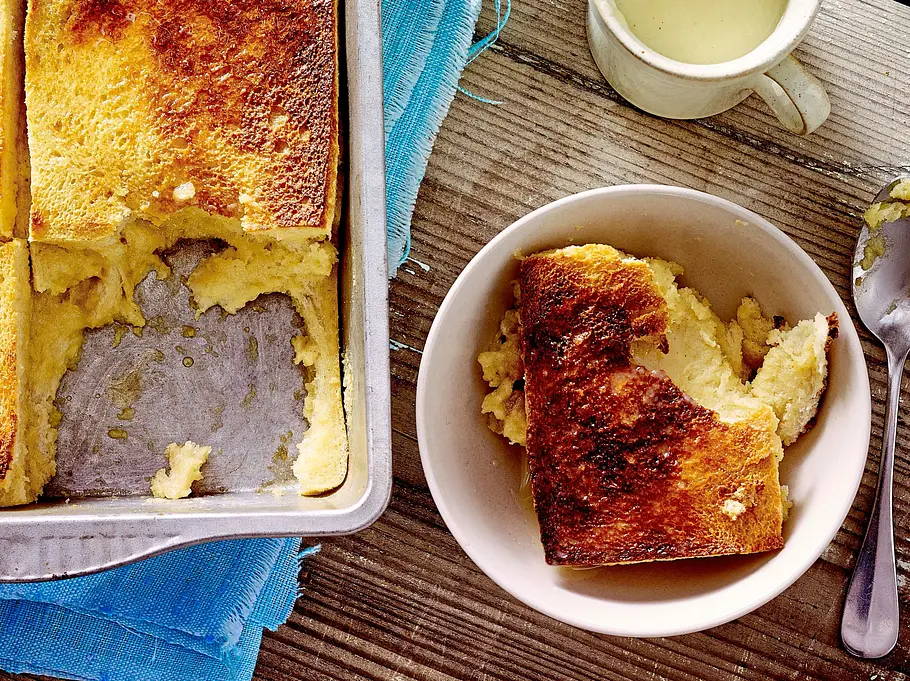 Goldbraun gebackener French-Toast-Auflauf in einer rechteckigen Form, mit knuspriger Oberfläche und saftiger Krume. Daneben ein Kännchen Vanillesoße und ein Stück des Auflaufs in einer Schale, serviert auf einem Holztisch mit Pfannenwender und Tuch.