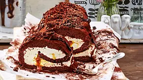 Schokoladige Biskuitrolle mit cremiger Sahnefüllung und Karamellkern, mit Schokoraspeln bestreut, auf Backpapier und einem weißen Holzbrett serviert. - Foto: LECKER @ Bauer Media Group Schokoladige Biskuitrolle mit cremiger Sahnefüllung und Karamellkern, mit Schokoraspeln bestreut, auf Backpapier und einem weißen Holzbrett serviert. - Foto: LECKER @ Bauer Media Group