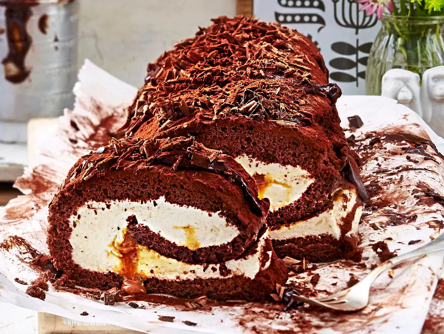 Schokoladige Biskuitrolle mit cremiger Sahnefüllung und Karamellkern, mit Schokoraspeln bestreut, auf Backpapier und einem weißen Holzbrett serviert.