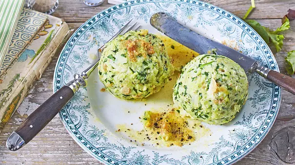 Zwei große Spinat-Ricotta-Klöße auf einem verzierten Teller, bestreut mit etwas Pfeffer und Butter, dazu Besteck und eine leicht strukturierte Holzunterlage. - Foto: LECKER @ Bauer Media Group