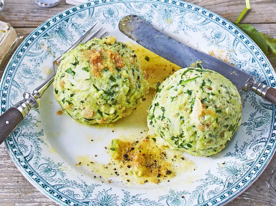 Spinat-Ricotta-Klöße Zwei große Spinat-Ricotta-Klöße auf einem verzierten Teller, bestreut mit etwas Pfeffer und Butter, dazu Besteck und eine leicht strukturierte Holzunterlage.
