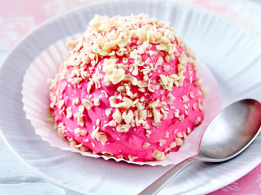 Eine kleine, kuppelförmige Pavlova in leuchtendem Rosa, umhüllt von einer Creme und bestreut mit gehackten weißen Nüssen, auf einem weißen Teller mit Papierförmchen und Löffel, dekoriert mit rosa Serviette.