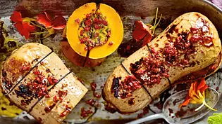 Ein goldgelb gebackener Butternut-Kürbis, prall gefüllt mit cremigem Hummus und knusprigem Topping aus Kräutern und Gewürzen, stilvoll auf einer Metallform angerichtet und mit essbaren Blüten garniert. - Foto: LECKER @ Bauer Media Group