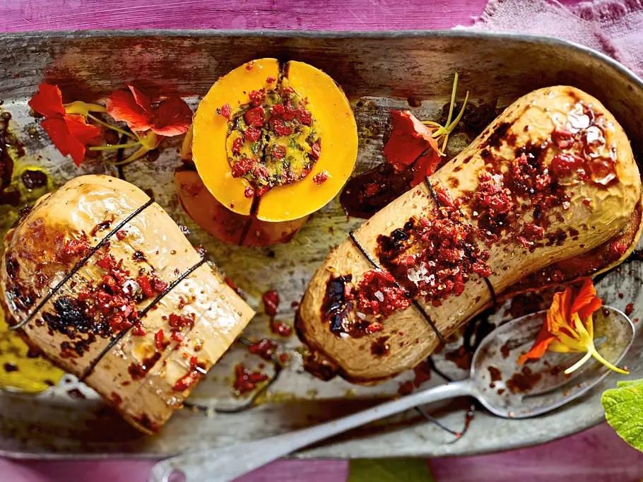 Ein goldgelb gebackener Butternut-Kürbis, prall gefüllt mit cremigem Hummus und knusprigem Topping aus Kräutern und Gewürzen, stilvoll auf einer Metallform angerichtet und mit essbaren Blüten garniert.