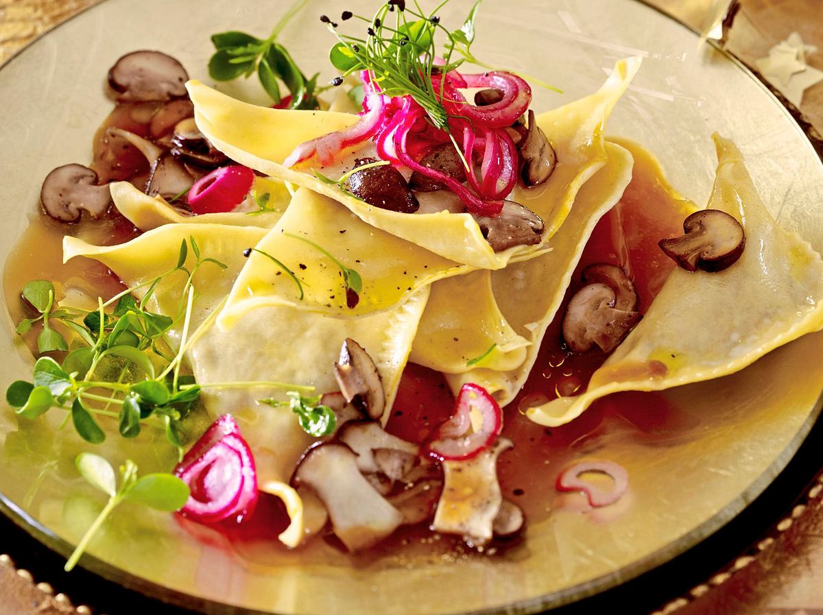 Ravioli mit Pilzfüllung, serviert in aromatischer Brühe, garniert mit eingelegten roten Zwiebeln, frischen Kräutern und gebratenen Pilzscheiben.