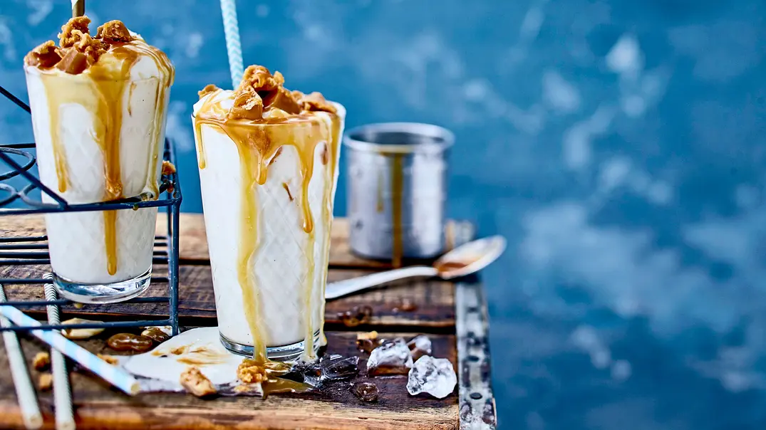 Cremiger Vanille-Milchshake in hohen Gläsern, großzügig mit goldbrauner Karamellsoße übergossen, die am Rand hinabläuft. Garniert mit Karamellstückchen und serviert mit Strohhalm auf einem rustikalen Holzbrett, daneben Eiswürfel und Löffel. - Foto: LECKER @ Bauer Media Group