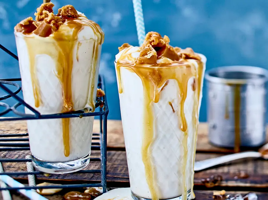 Cremiger Vanille-Milchshake in hohen Gläsern, großzügig mit goldbrauner Karamellsoße übergossen, die am Rand hinabläuft. Garniert mit Karamellstückchen und serviert mit Strohhalm auf einem rustikalen Holzbrett, daneben Eiswürfel und Löffel.
