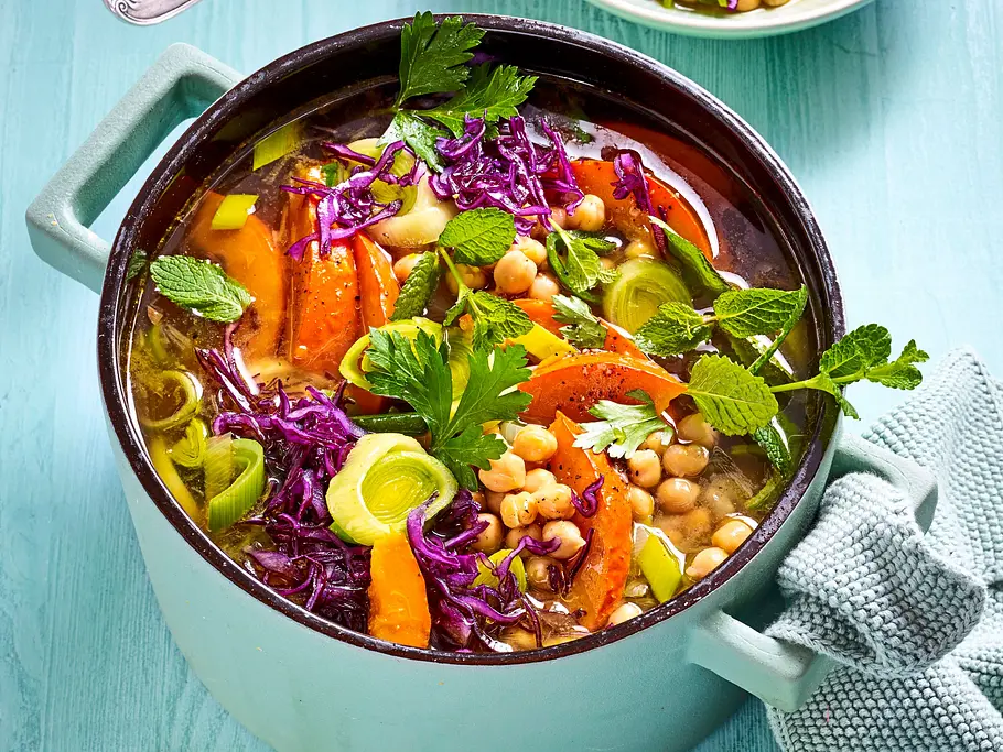 Low-Carb-Eintopf mit Kohl und Kürbis Bunter Low-Carb-Eintopf mit Kürbis, Rotkohl, Lauch, Kichererbsen und frischen Kräutern in einem hellgrünen Topf auf einem blaugrünen Holztisch.