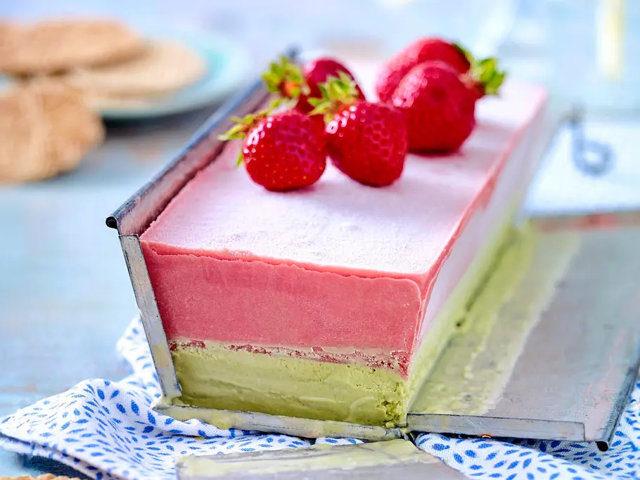 Erdbeer-Pistazien-Parfait Rechteckiges Erdbeer-Pistazien-Parfait mit zwei farbigen Schichten, belegt mit frischen Erdbeeren, auf einem hellen Dessertteller und einem gemusterten Tuch serviert.