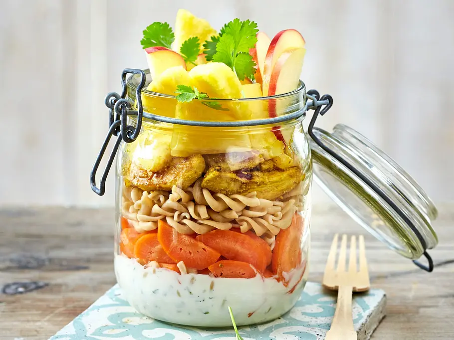 Curry-Nudelsalat zum Mitnehmen Ein großes Einmachglas mit einem Schichtsalat aus Joghurt-Dressing, Möhrenscheiben, Fusilli, Ananas und Äpfeln.