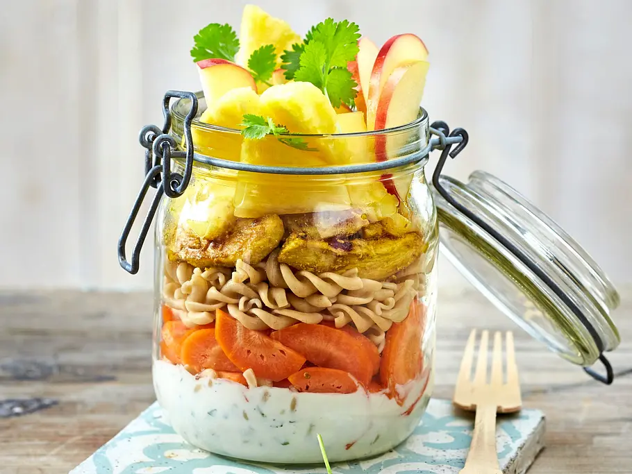 Curry-Nudelsalat im Einmachglas, geschichtet mit Joghurt-Dressing, Karotten, Vollkornnudeln, gebratenem Hühnchen, Ananas, Apfelscheiben und frischem Koriander, auf einer dekorativen Kachel.