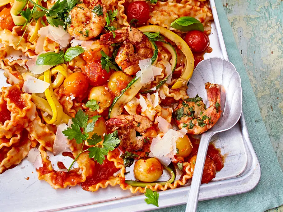 Mafaldine mit Garnelen Mafaldine-Nudeln mit gebratenen Garnelen, bunten Cherrytomaten, Zucchinistreifen, frischen Kräutern und Parmesanspänen auf einem silbernen Backblech angerichtet.