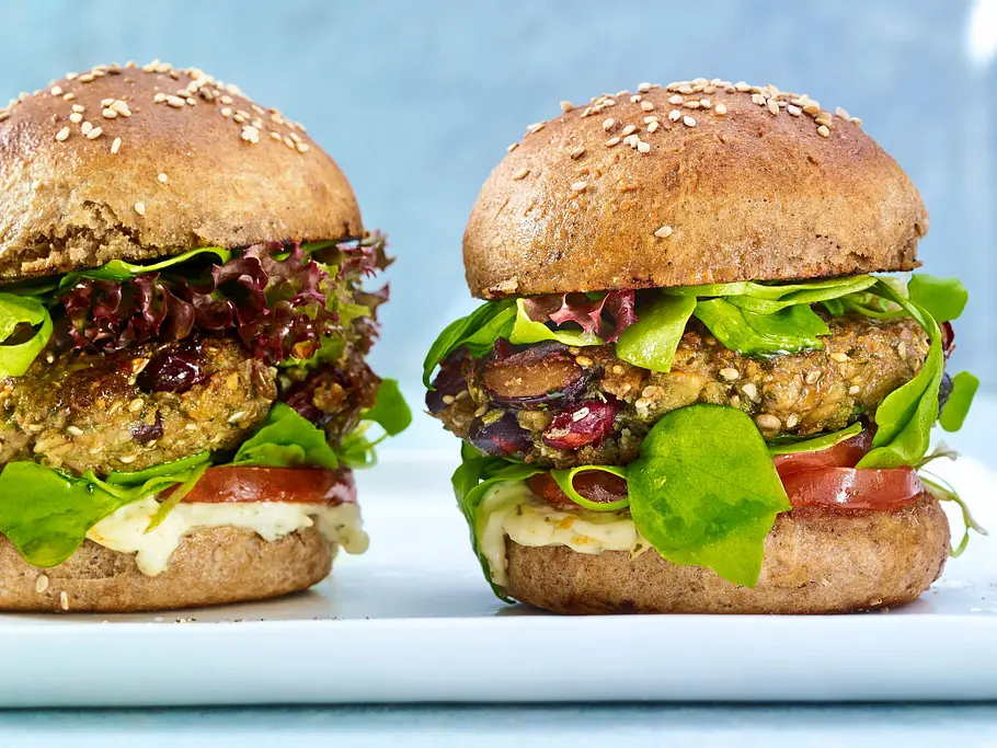 Zwei Grünkern-Burger mit Vollkornbrötchen, Salat, Tomaten, frischen Kräutern und cremigem Dip, auf einer rechteckigen weißen Platte.