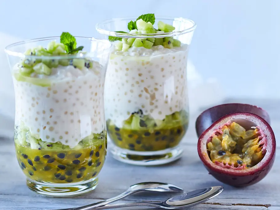 Zwei Gläser mit Kokos-Tapioka-Pudding, geschichtet mit Passionsfrucht und Gurkenstückchen, garniert mit Minze, daneben eine aufgeschnittene Passionsfrucht auf einem hellen Holztisch.