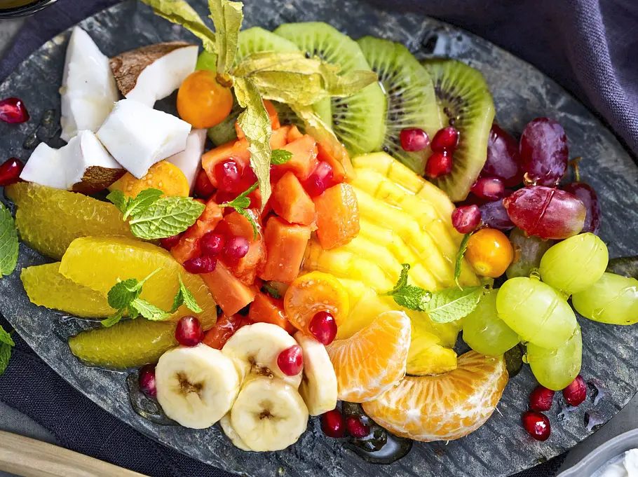 Tropen-Obstsalat mit Crema di Coco Rezept Bunter Tropen-Obstsalat mit frischer Mango, Kiwi, Banane, Papaya, Trauben, Orangenfilets, Kokosnussstücken und Granatapfelkernen, garniert mit Minze, auf einem dunklen Stein-Teller angerichtet und mit cremiger Crema di Coco in einer kleinen Schale serviert.