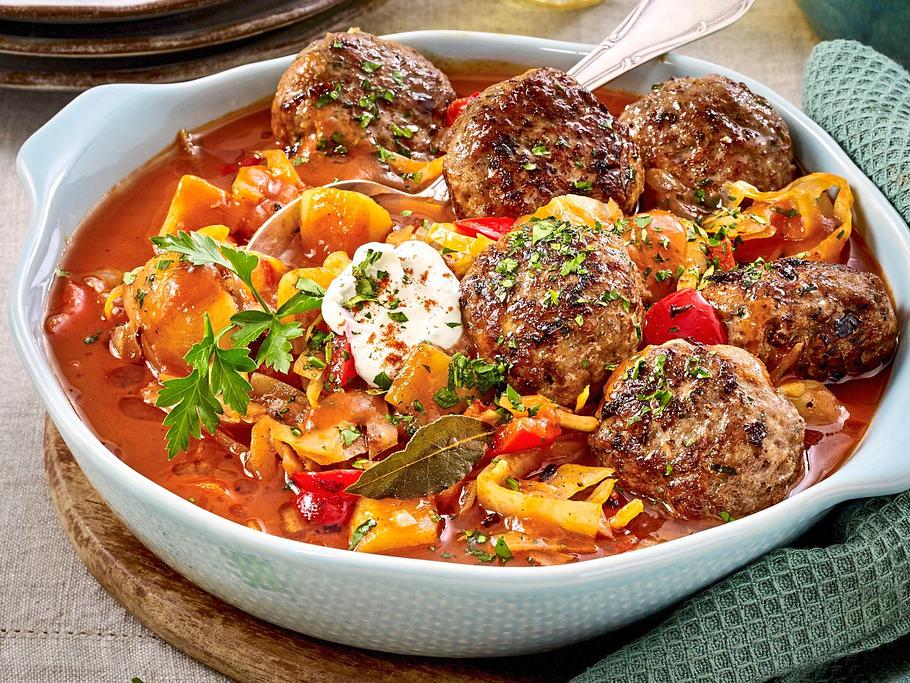 Deftiger Eintopf aus Kartoffeln, Weißkohl und Paprika in tomatiger Sauce, dazu gebratene Frikadellen und ein Klecks Sauerrahm, serviert in einer Auflaufform.