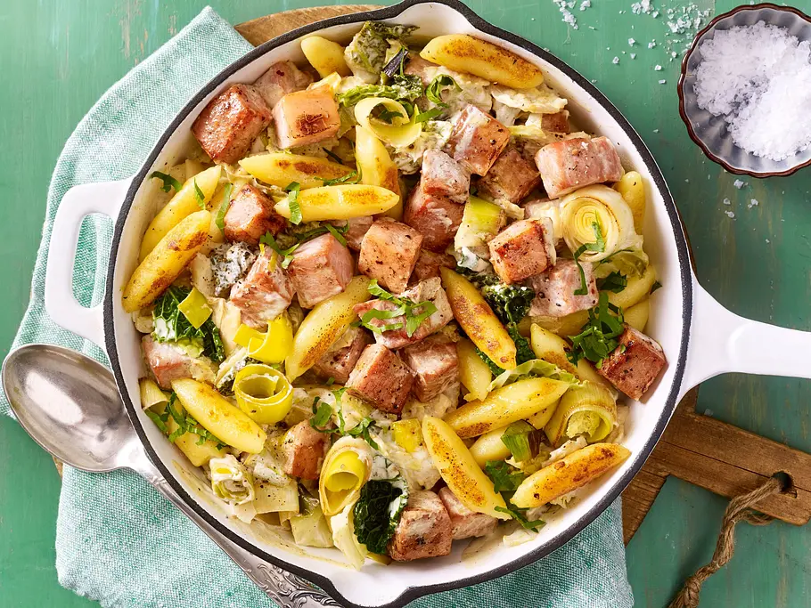 Gebratene Schupfnudeln mit Kasselerwürfeln, Lauch und Kräutern in einer weißen Pfanne auf einem grünen Tuch.