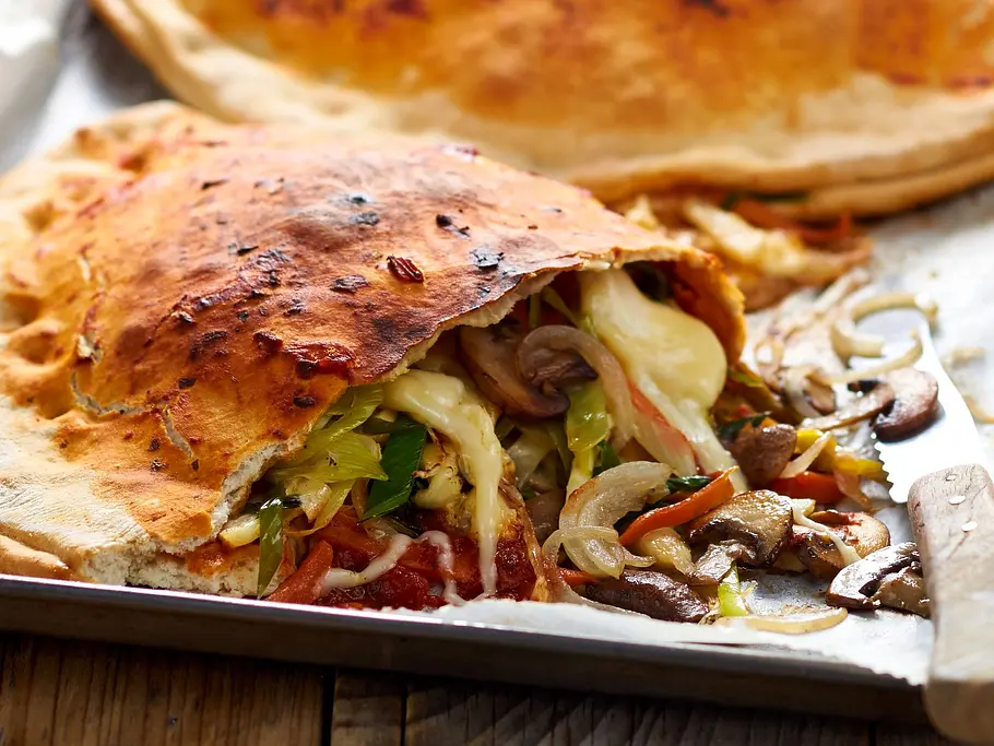 Goldbraune, knusprig gebackene Gemüse-Calzone, gefüllt mit geschmolzenem Käse, sautierten Pilzen, Lauch, Karotten und weiteren bunten Gemüsestreifen, serviert auf einem Backblech mit Backpapier.