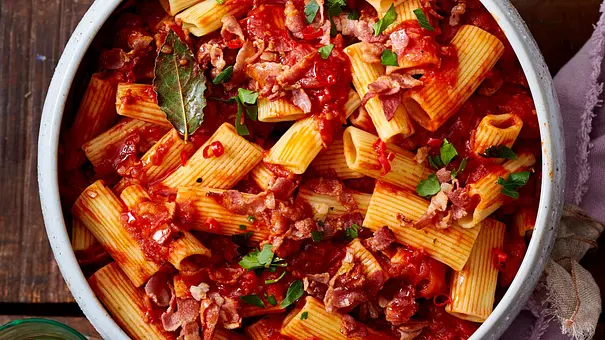 Rigatoni-Nudeln in leuchtend roter Tomatensoße mit Pancetta, garniert mit Petersilie, serviert in einer weißen Keramikschüssel auf einem rustikalen Holztisch. - Foto: LECKER @ Bauer Media Group
