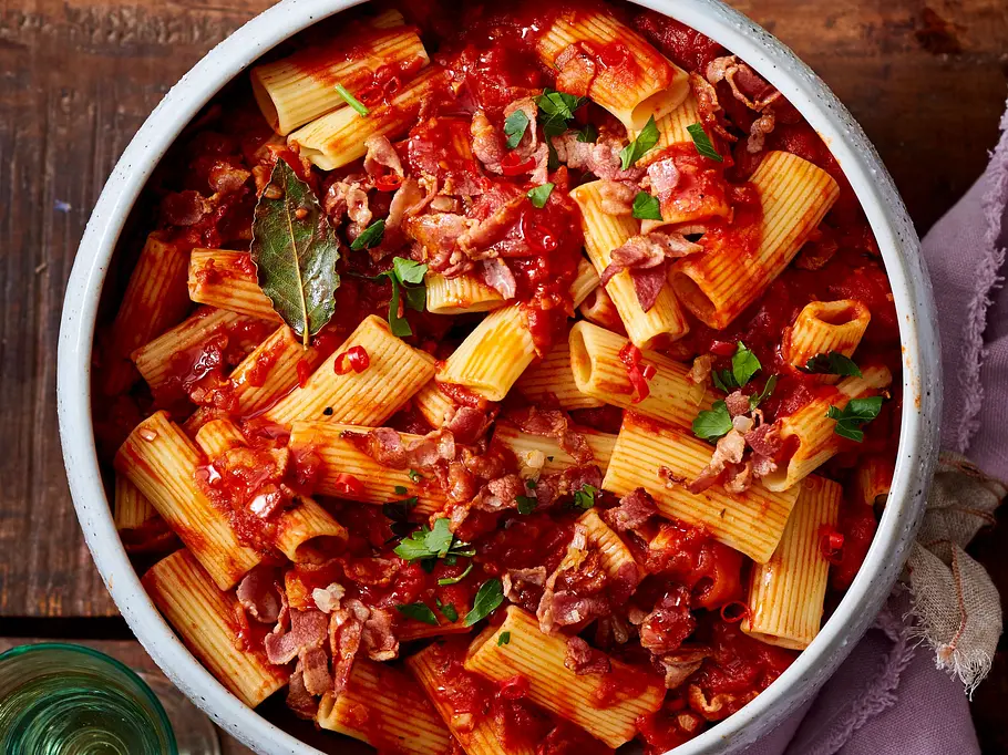Rigatoni in Tomaten-Pancetta-Soße Rigatoni-Nudeln in leuchtend roter Tomatensoße mit Pancetta, garniert mit Petersilie, serviert in einer weißen Keramikschüssel auf einem rustikalen Holztisch.