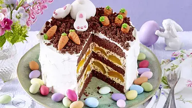 Stracciatella-Torte mit lockeren Schokoböden, cremiger Füllung und Sahne umhüllt, dekoriert mit Schokoraspeln, Marzipanmöhren, kleinen Osterhasen und bunten Zuckereiern, serviert auf einer grünen Platte. - Foto: LECKER @ Bauer Media Group