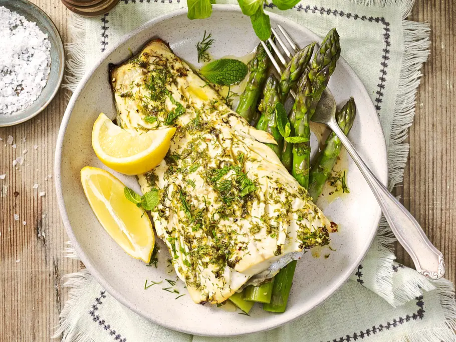 Schneller Ofenfisch mit grünem Spargel, zart gegartes Fischfilet mit frischen Kräutern bestreut, serviert mit Zitronenspalten auf einem hellen Teller, angerichtet auf einem Leinentuch mit grobem Meersalz und Pfeffermühle daneben.