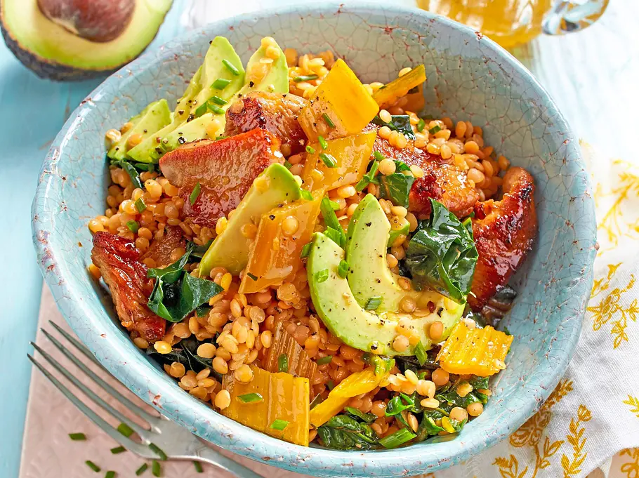 Warmer Linsensalat mit roten Linsen, Avocadostreifen, gelbem Gemüse und gebratenen Tomaten in einer türkisfarbenen Keramikschüssel.