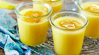 Gelbe Melonen-Gazpacho mit einem Tupfer Honig, serviert in kleinen Gläsern auf einem rustikalen Holzbrett und einem zarten, geblümten Tuch. - Foto: LECKER @ Bauer Media Group
