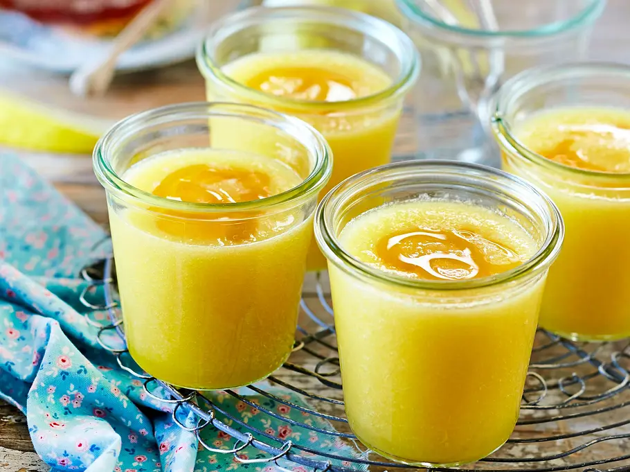 Gelbe Melonen-Gazpacho mit einem Tupfer Honig, serviert in kleinen Gläsern auf einem rustikalen Holzbrett und einem zarten, geblümten Tuch.