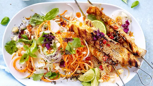 Zwei gegrillte Puten-Spieße auf einem weißen ovalen Teller, serviert mit bunten Gemüsenudeln aus Karotten und Zucchini, frischen Kräutern und Limettenspalten. Daneben ein Schälchen mit Dip. - Foto: LECKER @ Bauer Media Group