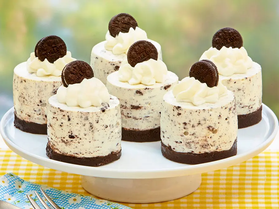 Frozen Stracciatella-Törtchen mit Oreos Sechs kleine, gefrorene Stracciatella-Törtchen mit Schokoladenkeksboden und Sahnehaube, jeweils mit einem Schokokeks dekoriert, auf einer weißen Tortenplatte, serviert auf einer gelb karierten Tischdecke.