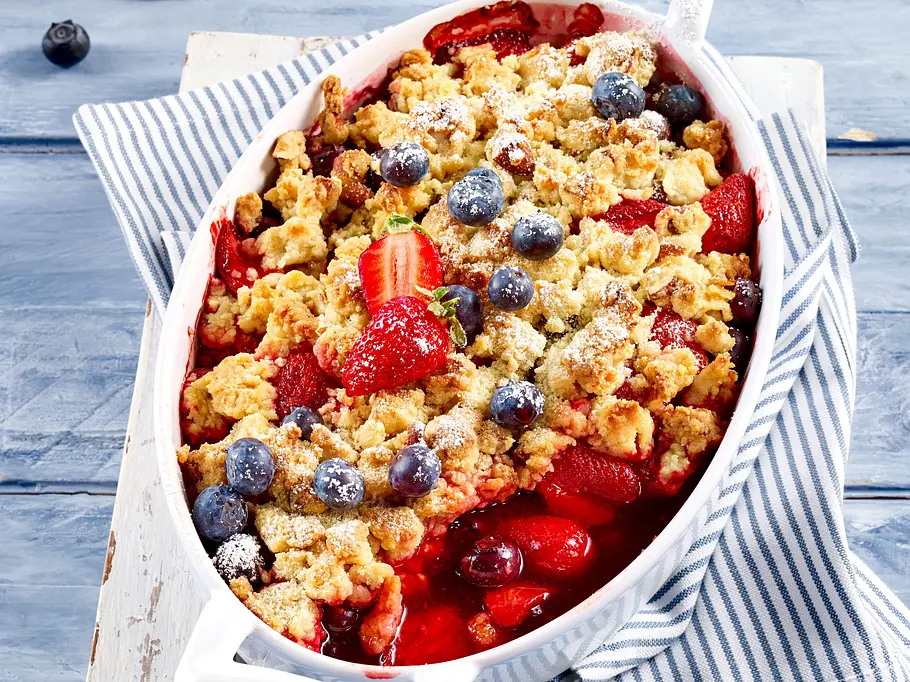 Beeren-Crumble Beeren-Crumble mit knusprigen Streuseln und frischen Erdbeeren und Heidelbeeren in einer weißen Auflaufform auf einem Holzbrett und gestreifter Stoffserviette.
